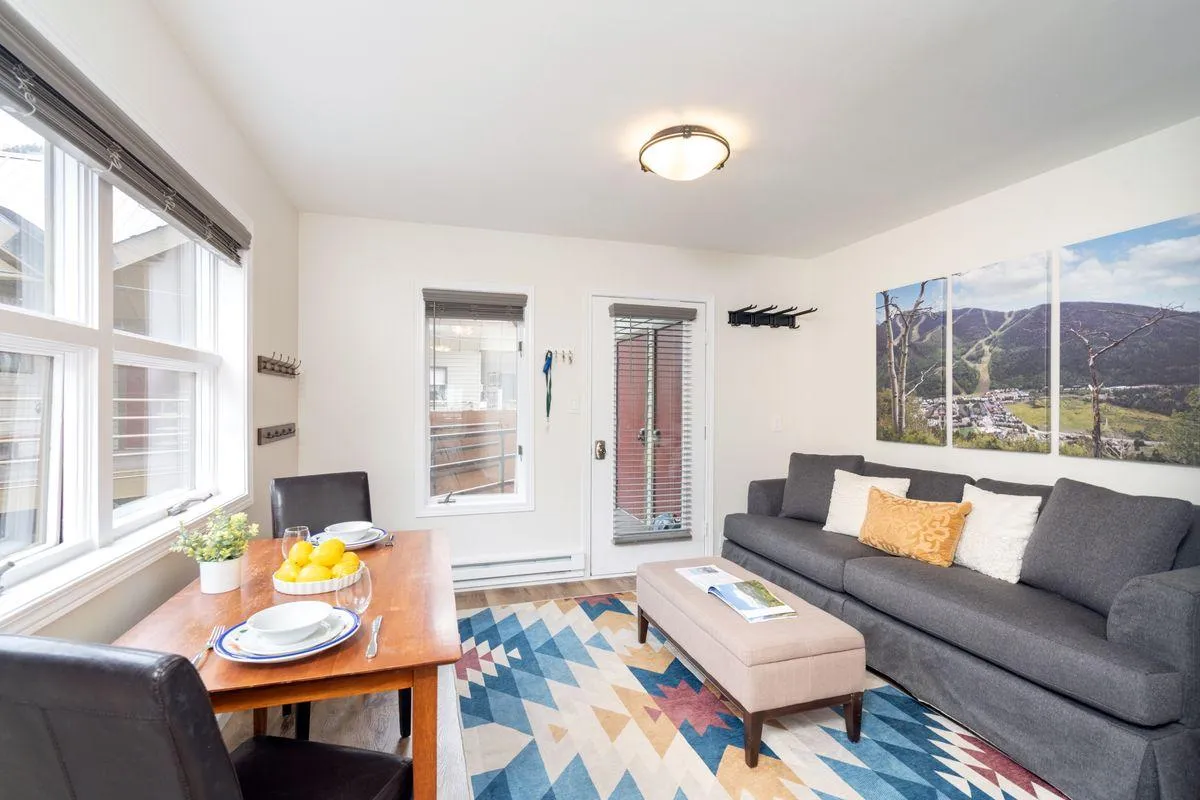 The hero image showcases a bright, welcoming living and dining area with a cozy gray sofa, a colorful area rug, a table set with a fresh fruit centerpiece, and a striking landscape artwork on the wall. Sunlight fills the space, creating an inviting atmosphere perfect for relaxing or gathering. The decor hints at mountain adventures outside while ensuring guests feel at home inside.