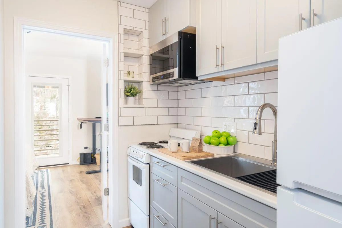 A bright, fresh kitchen space featuring a spacious countertop, green apples for a homey touch, crisp white cabinetry, and an open doorway leading into the living area. The layout is ideal for both casual dining and meal preparation, and the overall mood is inviting and modern.