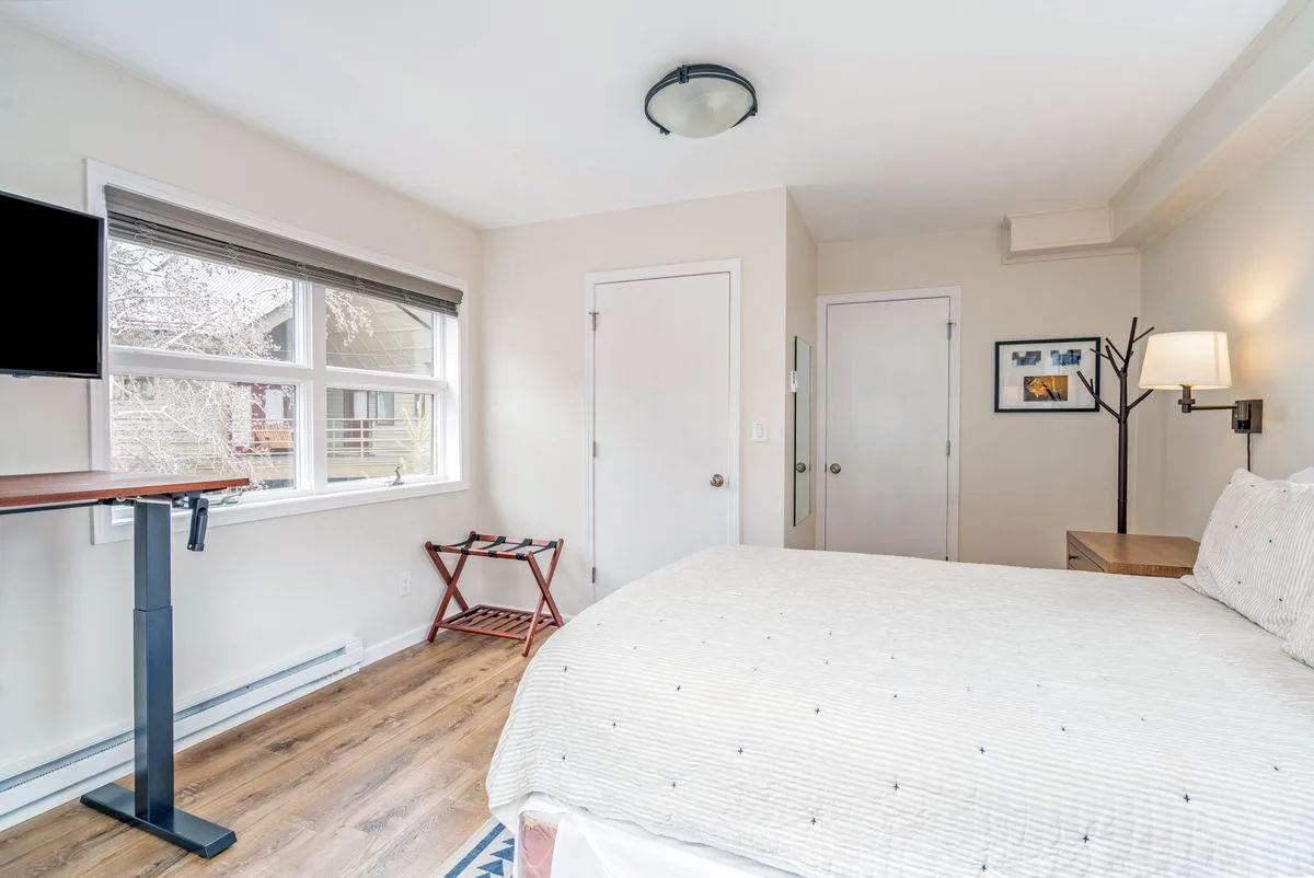 A cozy bedroom scene with a white-bedded queen bed, wall-mounted television, wooden side table, and a lamp by the window. Natural light fills the room, creating a calm, inviting atmosphere ideal for both couples and families seeking restful nights.