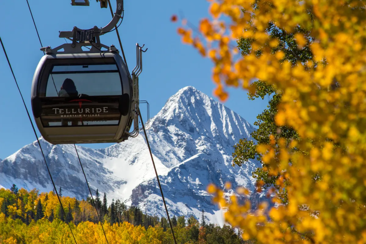 A gondola rises above colorful autumn foliage, framed by a snowy mountain in the background. The bright gondola car is prominent, with the grandeur of Telluride’s alpine scenery visible all around. The scene evokes a sense of adventure and exploration, hinting at sunrise or sunset rides.