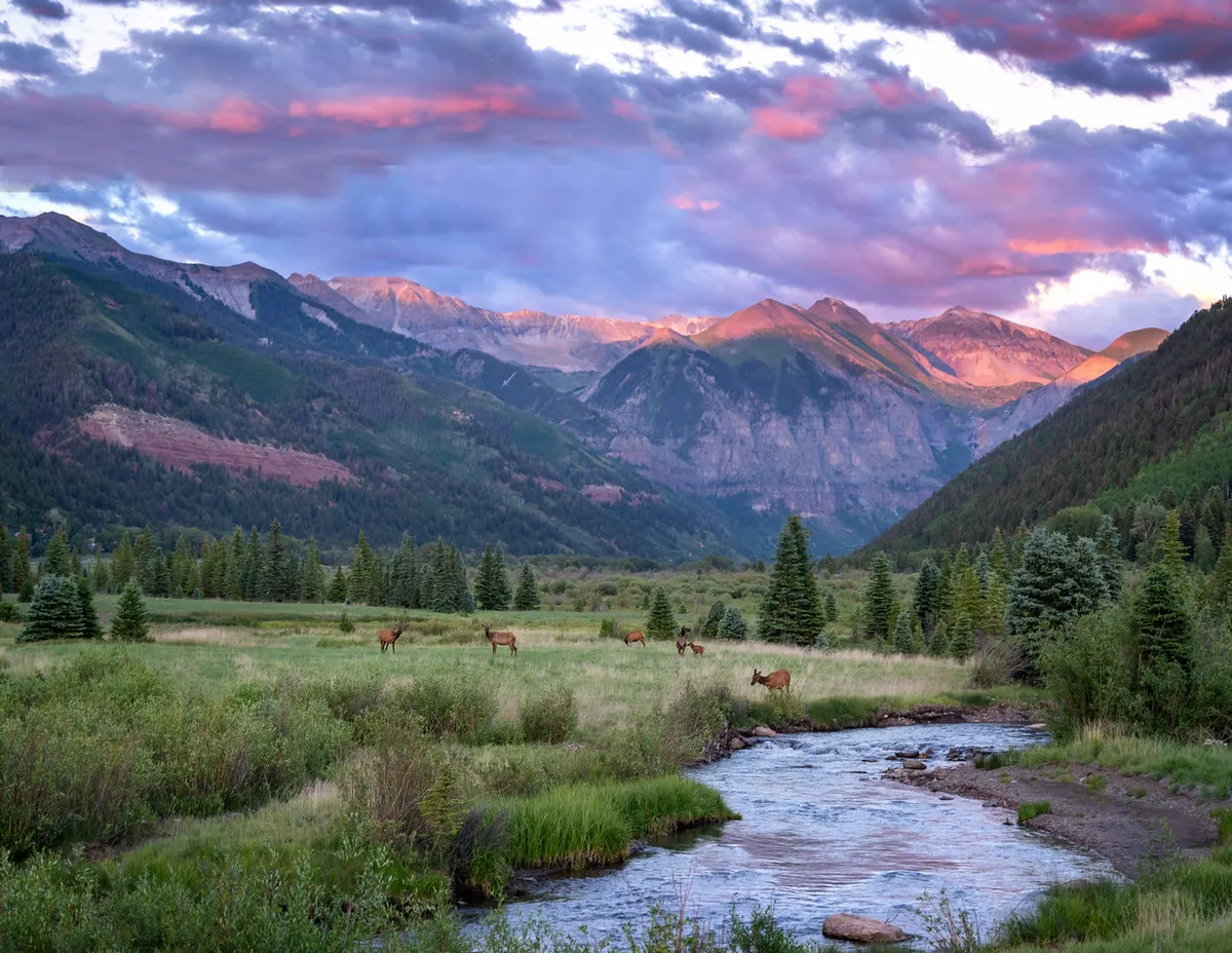 A serene riverside scene with a mountain backdrop, a gently flowing stream, and deer grazing along the bank. Wisps of clouds hover above, and the atmosphere is tranquil and inviting.