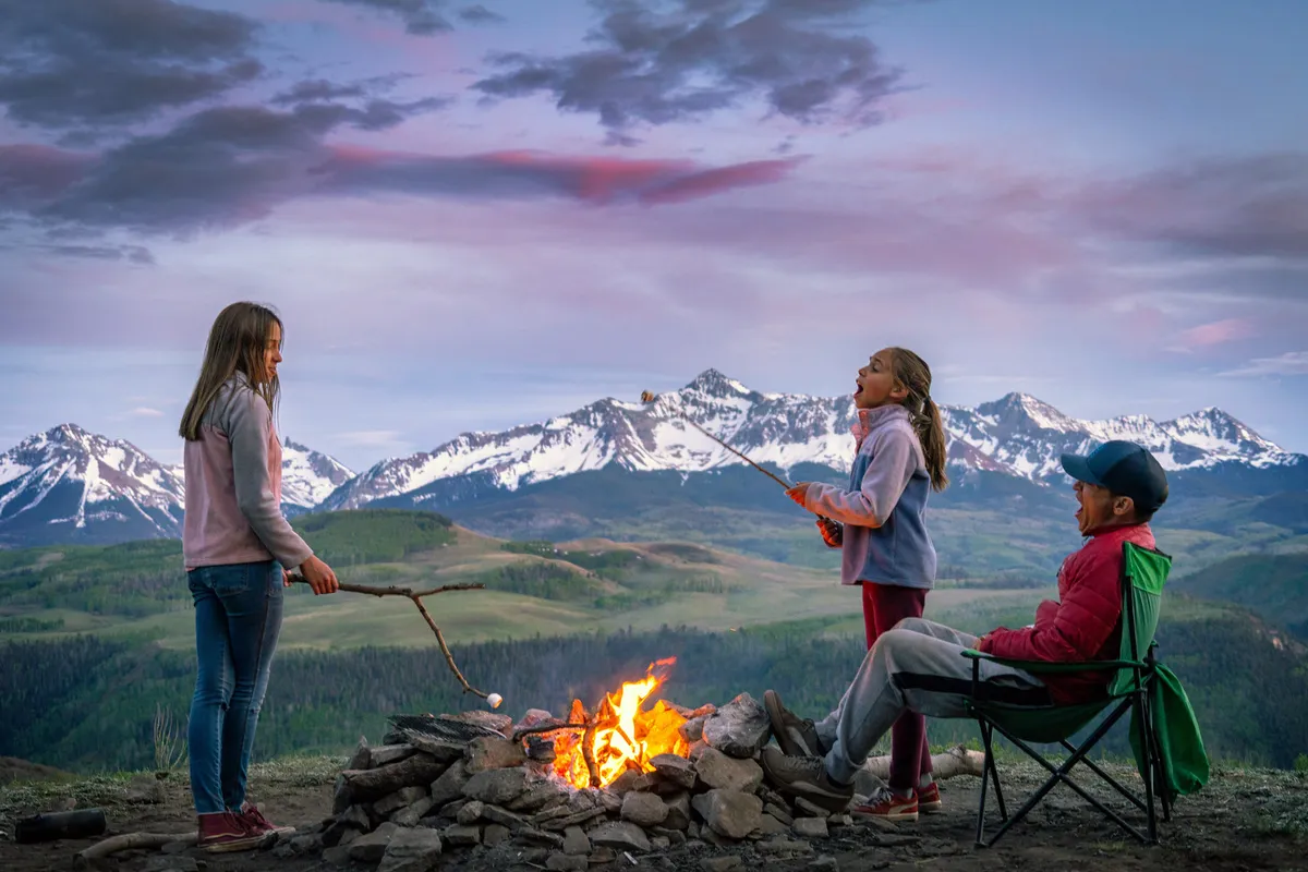 A joyful scene set by a campfire, with children roasting marshmallows, a man sitting in a chair, and a mountain landscape beyond. The image radiates familial warmth, togetherness, and fun, evoking playful and memorable moments families can create outdoors.