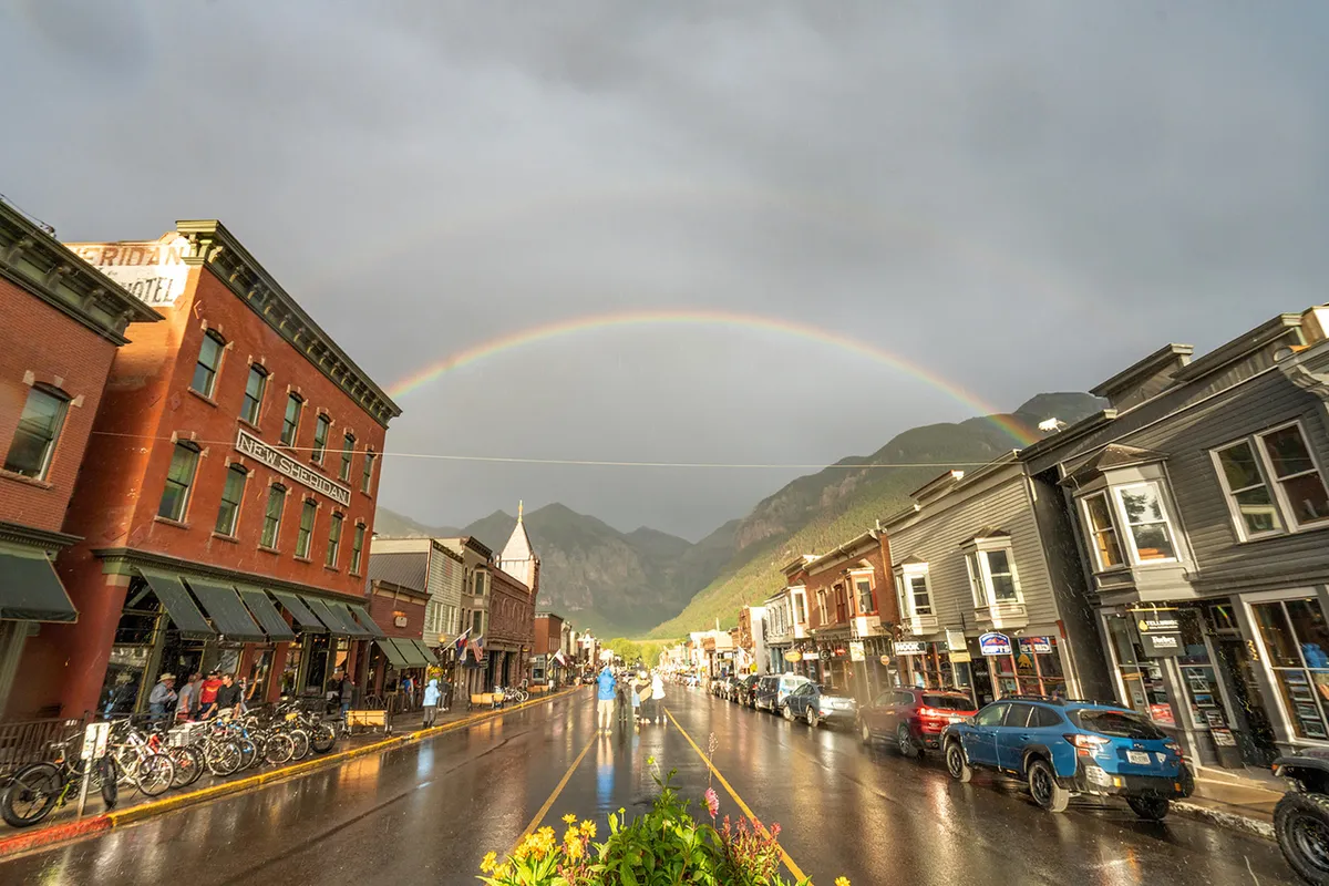 Historic, colorful downtown buildings line a wet street glistening after rain, with a rainbow arching overhead and mountain peaks in the distance. The scene suggests the charm of Telluride’s central area and its cozy, inviting ambiance. Cafés, shops, and restaurants are nestled along the street, ready for exploration and intimate dinners.
