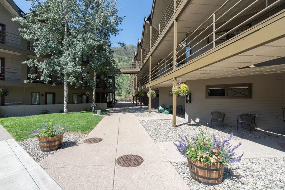 This exterior image shows a charming courtyard with flower planters, a winding pathway, balconies, and mature trees. The scene conveys tranquility and offers a glimpse into the outdoor communal areas that guests can enjoy, suggesting relaxation and outdoor socializing in a lush environment.
