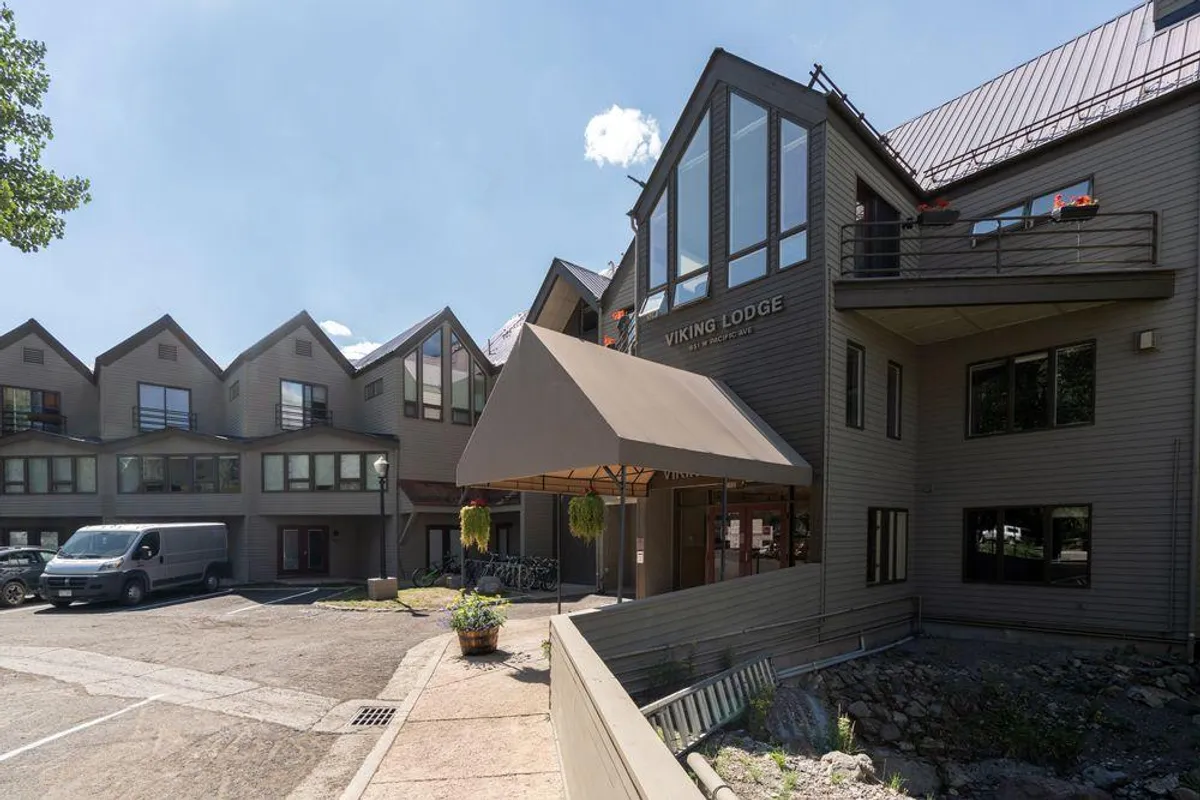 The image features the welcoming entrance of the Viking Lodge, highlighted by a prominent entrance awning, large windows, planters filled with greenery, and the lodge's sign. The scene is inviting, with a cozy and communal atmosphere suggesting arrival and a sense of place.