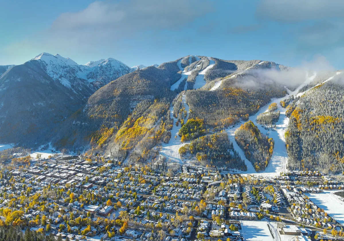 A panoramic view of snow-capped mountains, ski slopes, vibrant autumn trees, and a charming town nestled below. The mix of seasonal colors and visible town highlights the area's year-round appeal for both adventure and exploration.