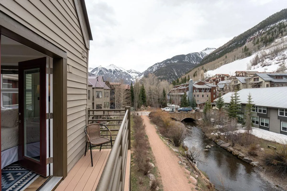 A peaceful river runs alongside a winding path, with a private balcony featuring a comfortable chair in the foreground. Snow-dusted mountains rise in the distance, and soft sunlight glows over the entire scene. The balcony overlooks the tranquil San Miguel River, connecting directly to the scenic surroundings and nearby town.