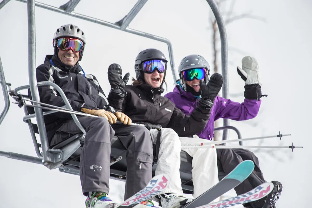 Three skiers ride a chairlift high above a snow-covered landscape, with ski equipment in hand and the wintry terrain stretching out beneath them. The image exudes the anticipation and excitement of a day on the slopes, while also highlighting the easy access to skiing and snowboarding.
