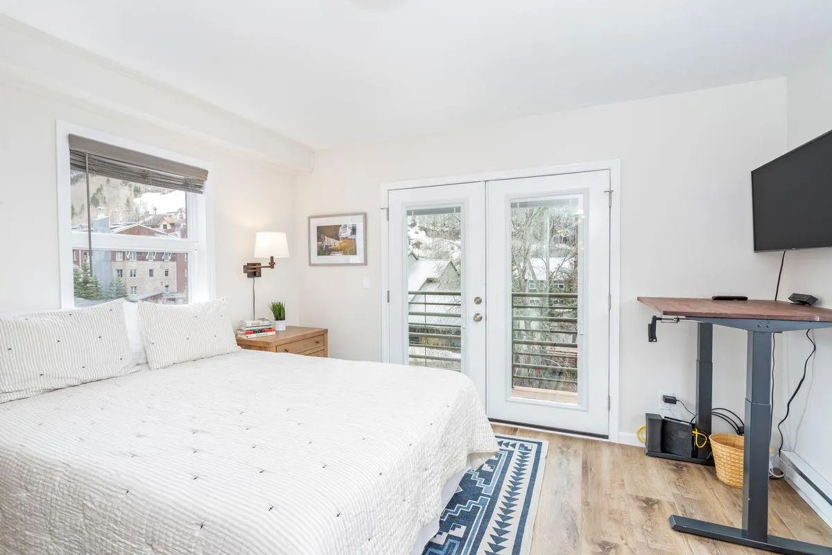 A serene, sunlit master bedroom featuring a plush queen bed positioned to capture natural light. French doors at the foot of the bed open to a private balcony, while a wall-mounted flat screen TV and elegant nightstand enhance comfort. The space exudes calm, inviting relaxation and offering a glimpse of outdoor tranquility.