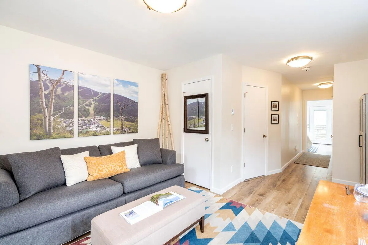 A welcoming corner of the living room with a plush sofa, coffee table, and mountain-themed wall art. The arrangement encourages relaxation and socializing, with a cozy, intimate feel complemented by natural light filtering through the windows.