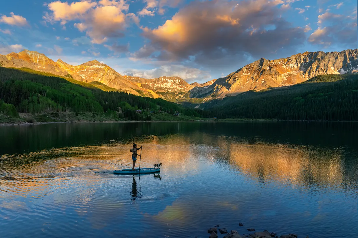 This tranquil exterior scene captures a paddleboarder and their dog gliding across a reflective body of water, framed by a stunning mountain range. The mood is peaceful, and the natural beauty encourages outdoor exploration and the enjoyment of Telluride’s riverside and trails.